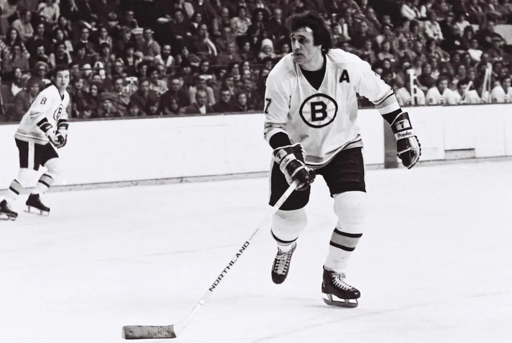 A hockey player wearing a Boston Bruins "B" jersey skillfully skates with the puck on the ice, an opponent shadowing him in the background, while an eager crowd watches from the stands.