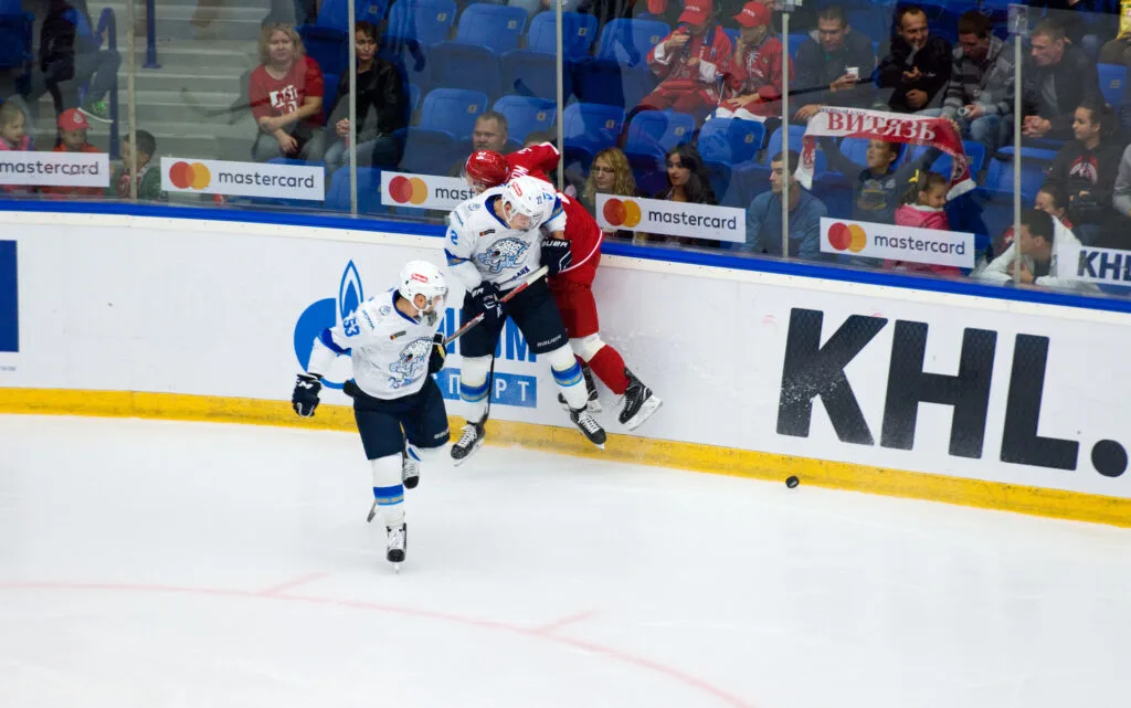 Two ice hockey players collide against the rink boards during a game, risking a boarding penalty as spectators watch intently in the background.