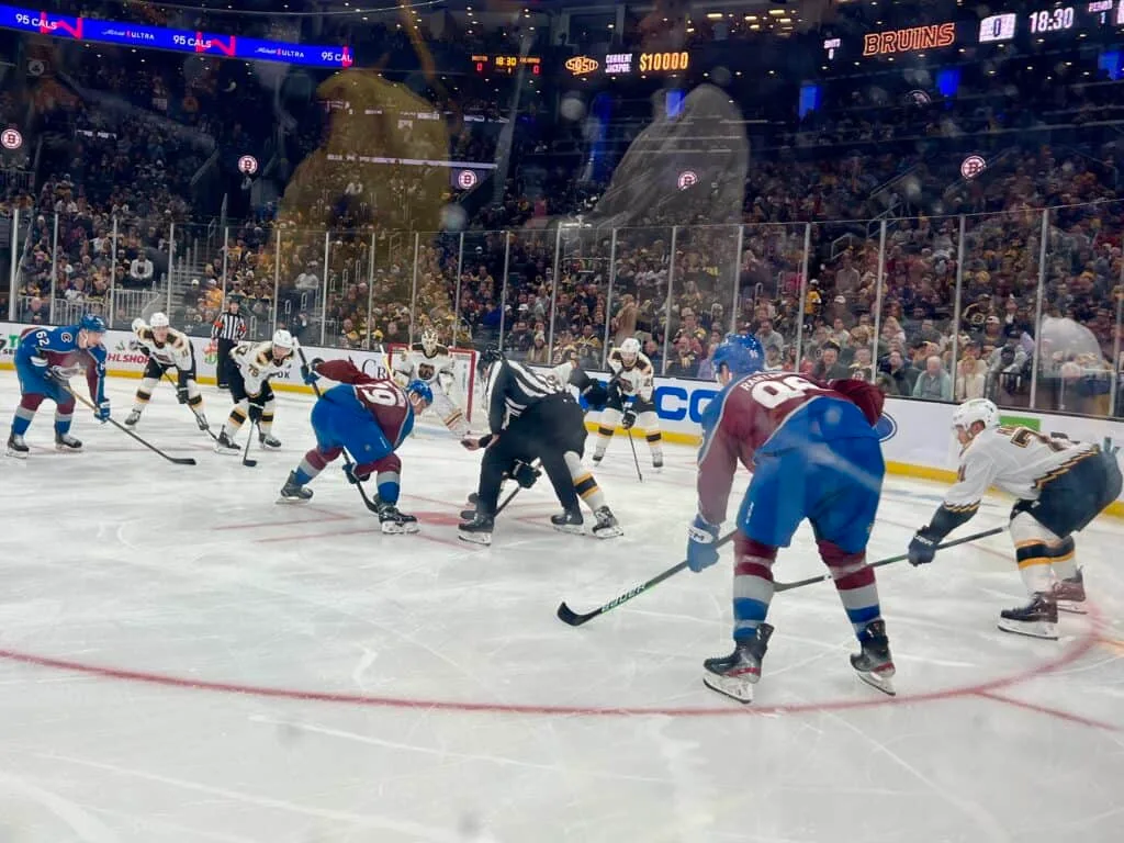 Colorado Avalanche hockey players from two teams face off on the ice rink during an NHL game, with referees nearby and a large crowd in the background.